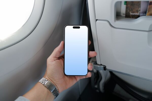 Hand holding an iPhone 15 Pro mockup with a blank screen inside a bright airplane cabin.